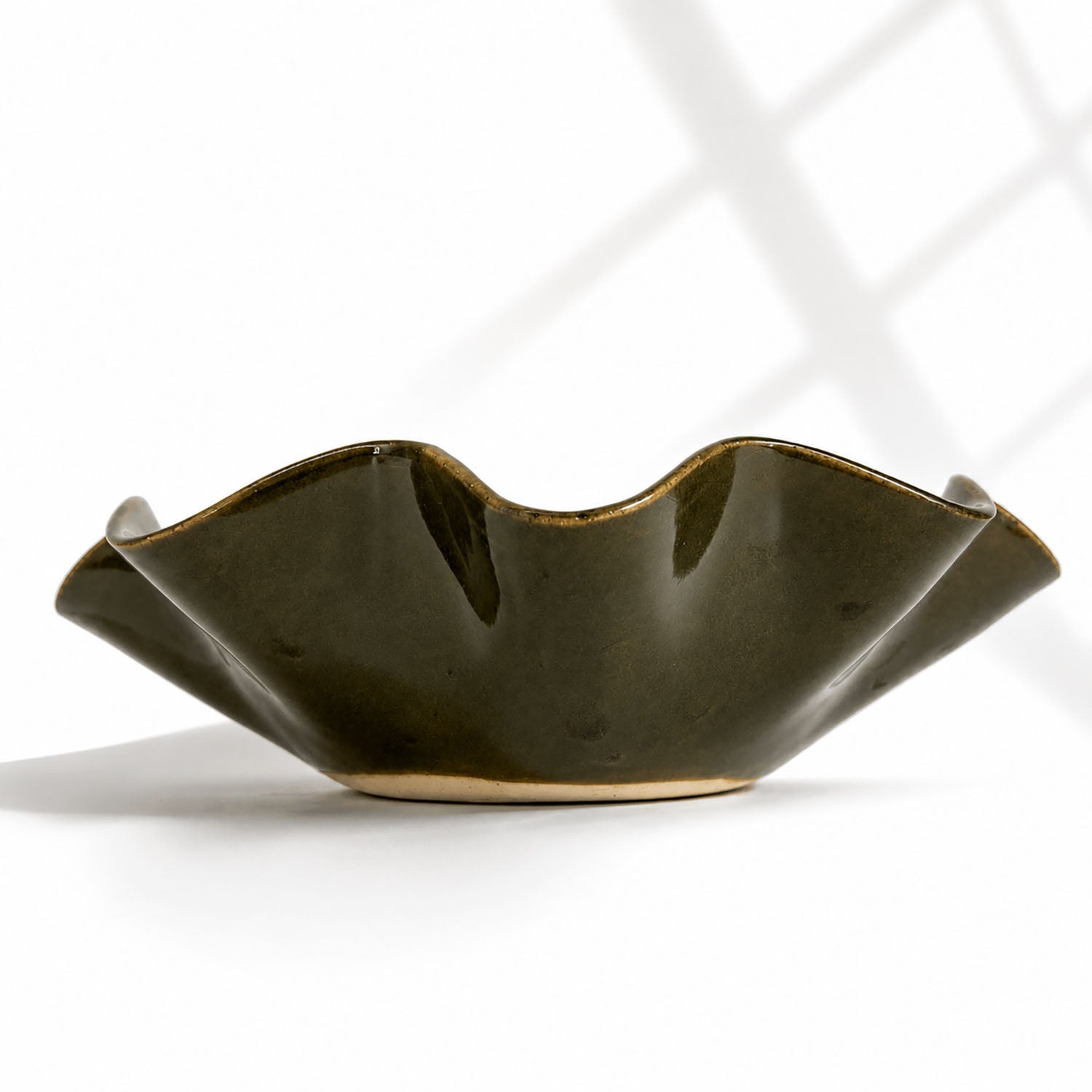 A dark olive green ceramic bowl by Polly Barton, with a wavy flared rim and a glossy finish, photographed on a white surface with soft window-light shadows.