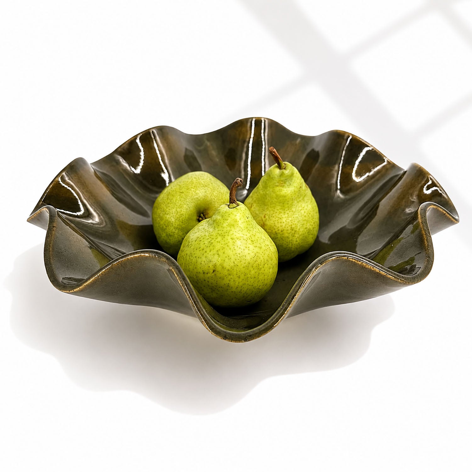 An oily Rockpool coloured wavy ceramic bowl with a glossy glaze, containing three green pears, set against a clean white background with soft window shadow patterns.