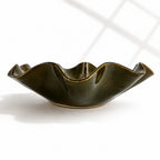 A dark olive green ceramic bowl with a wavy, flared rim, shown on a clean white background with soft window shadow patterns.