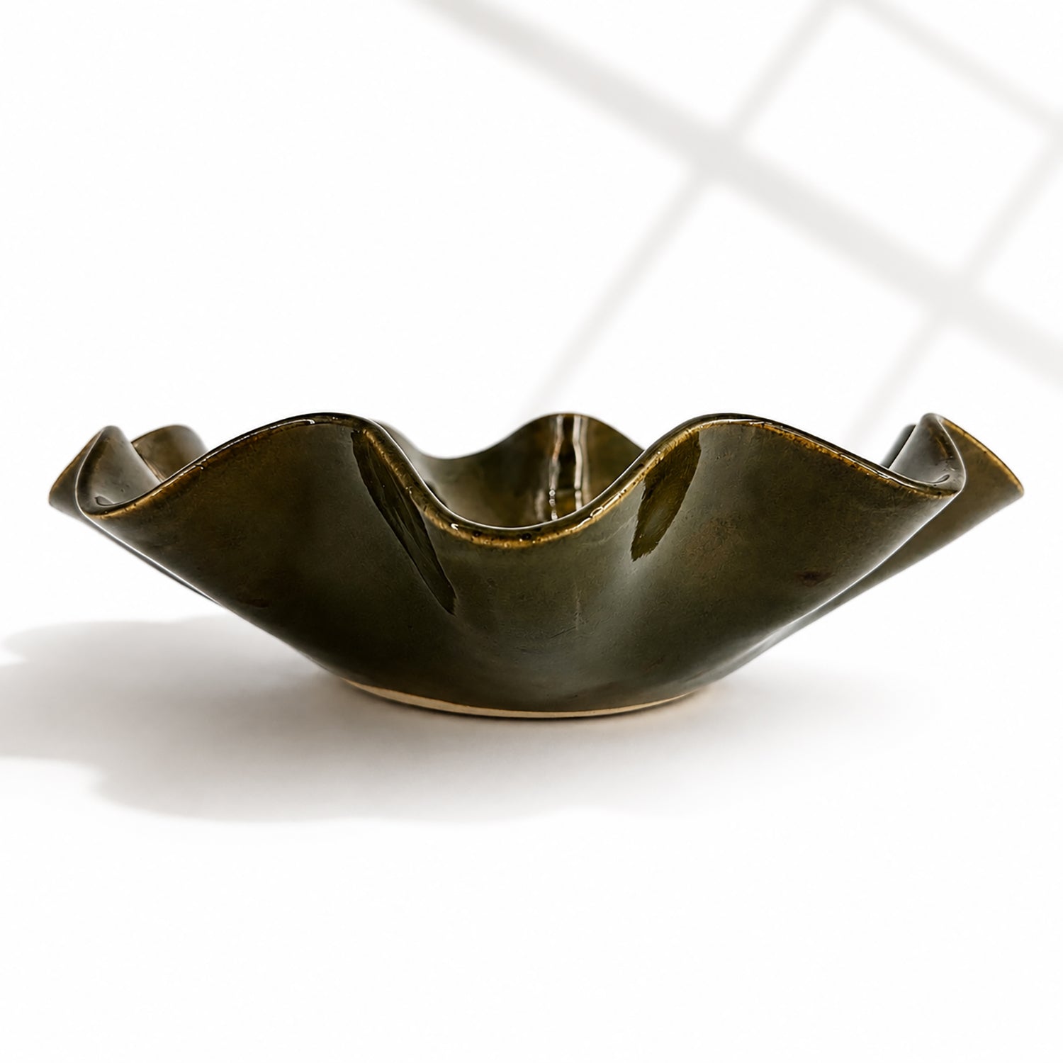 A dark olive green ceramic bowl with a wavy, flared rim, shown on a clean white background with soft window shadow patterns.