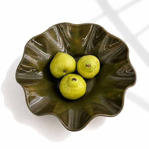 A wavy ceramic bowl with a glossy dark olive-green glaze, containing three green pears, photographed from above on a white background with soft window-light shadows.