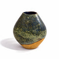A handmade ceramic vase with a rounded, bulbous shape, featuring a textured speckled glaze in olive green and dark blue-gray, with a contrasting rough, sandy gold base, set against a plain white background.