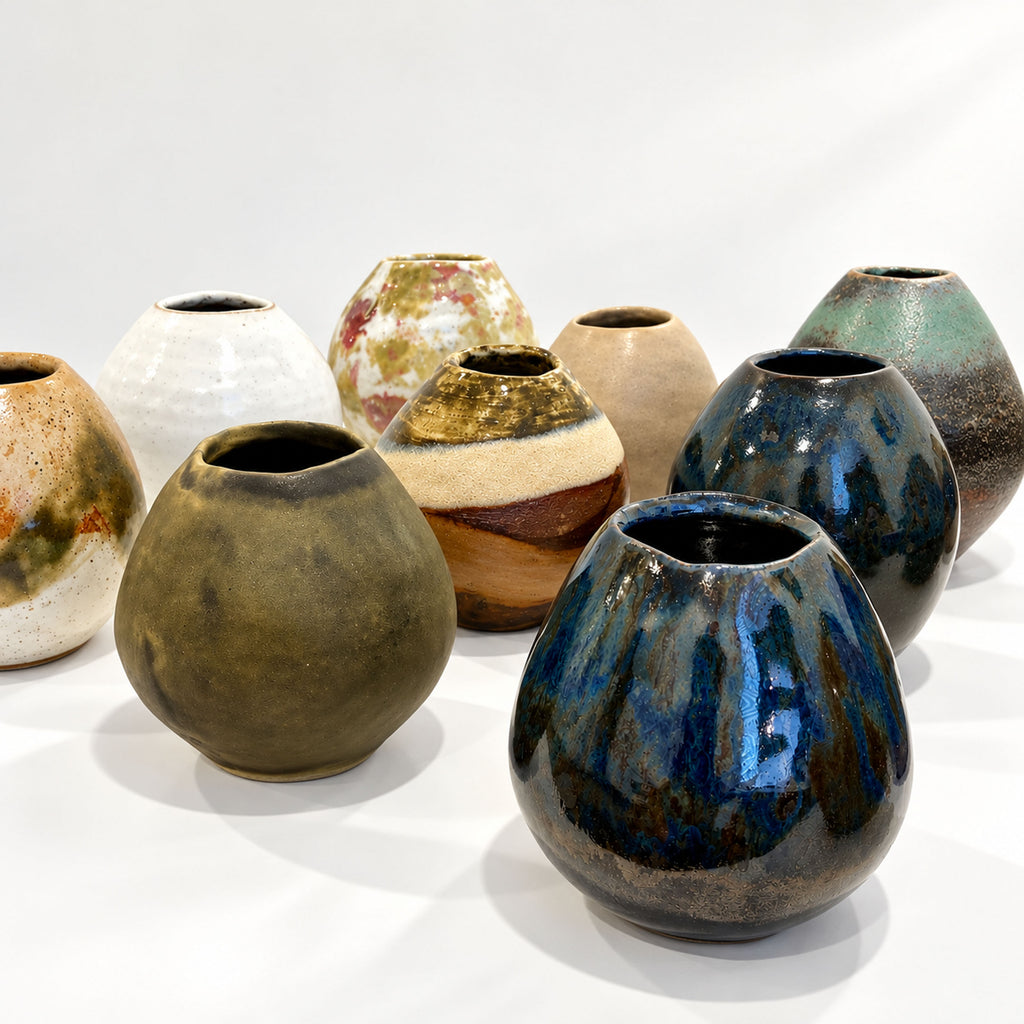 A group of handmade ceramic vases with diverse glazes and earthy tones, arranged on a white surface against a white background.